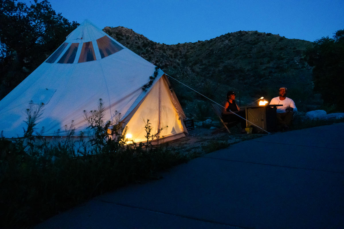 Man and woman outside in the desert sitting at a camp fire next to a large white tent, glamping