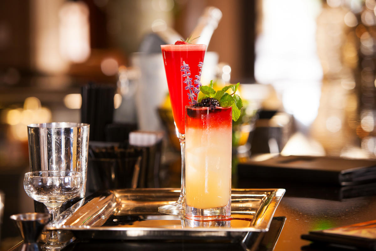 two cocktails — one bright red in a tall champagne glass, the other a gradient from red to a pale yellow liquid in a tall glass — sitting on a silver tray