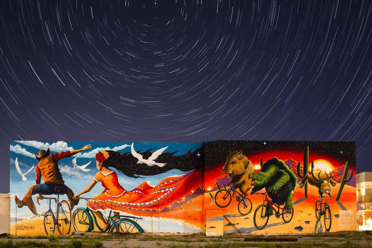 Colorful Mural spanning between two buildings. Male and Female on bikes riding into blue sky. Woman's dress flows behind and transform into sunset desert landscape. A javelina, tortoise, and jack rabbit on bikes follow behind couple