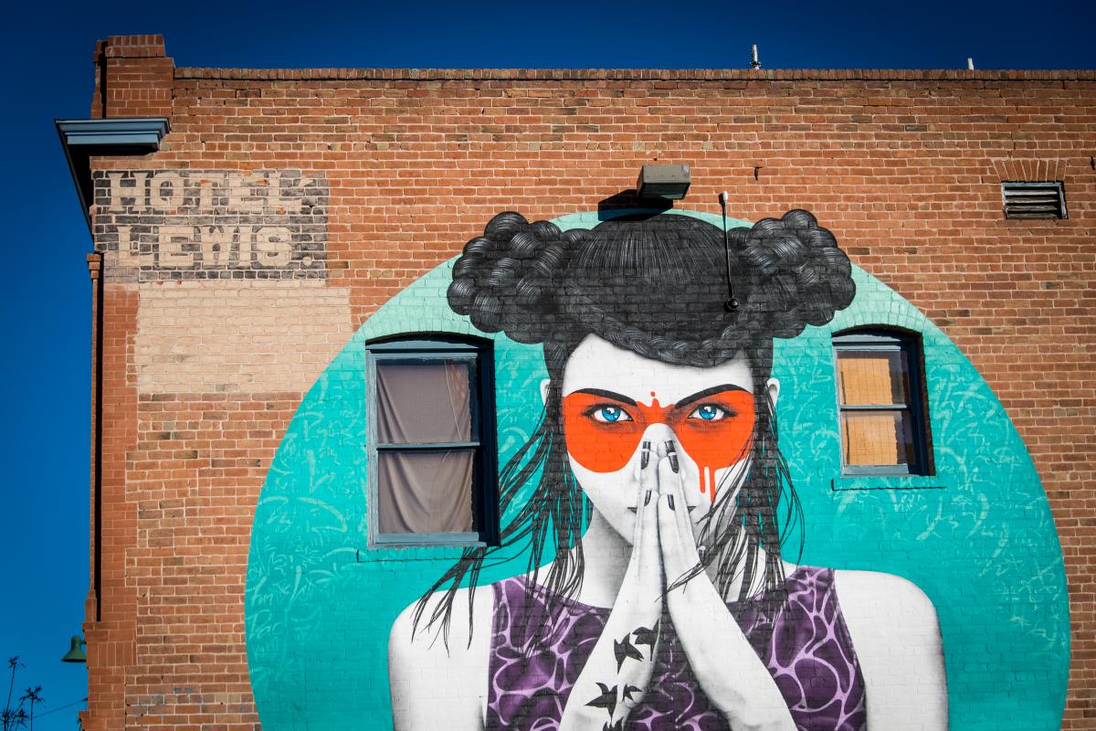 mural of a women posed with hands in front of face "prayer hands". red painted mask over her eyes. On her arm is a tattoo of several birds  and cursive text that reads "Forget me not"