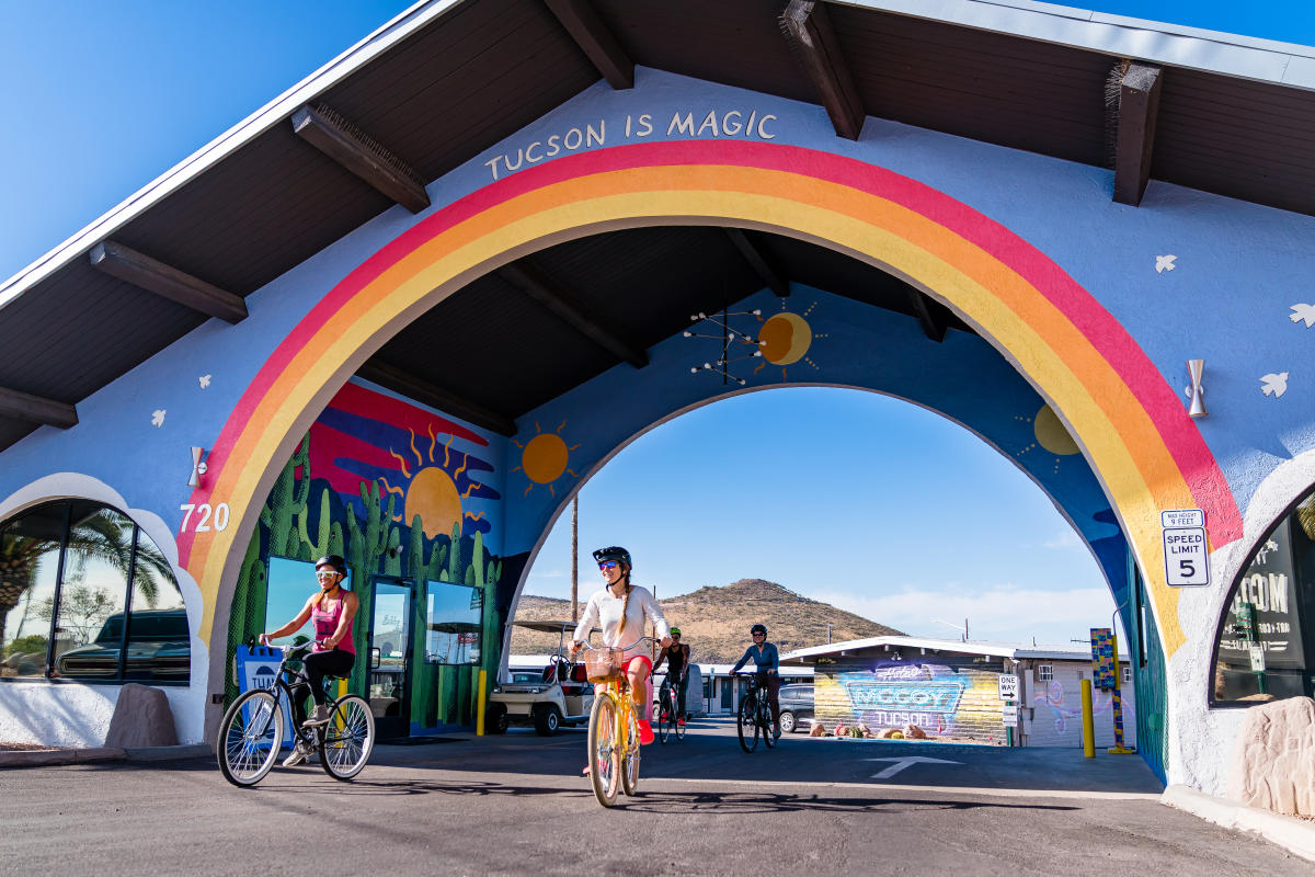 Group of friends riding bikes underneath Hotel McCoy's colorful, rainbow "Tucson is magic" mural