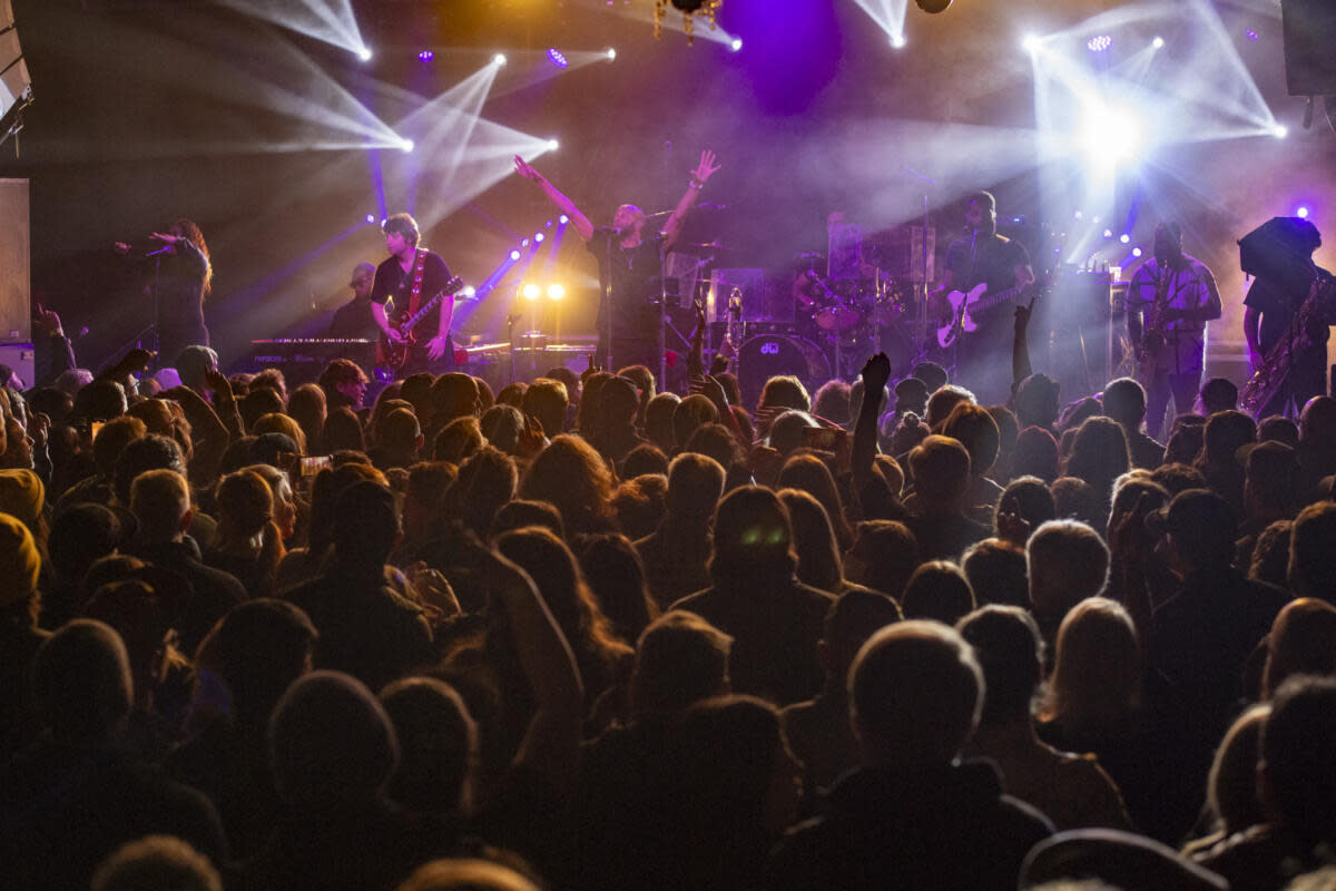 A band performing on stage with bright, colorful lights, while a large crowd watches, with their hands raised.
