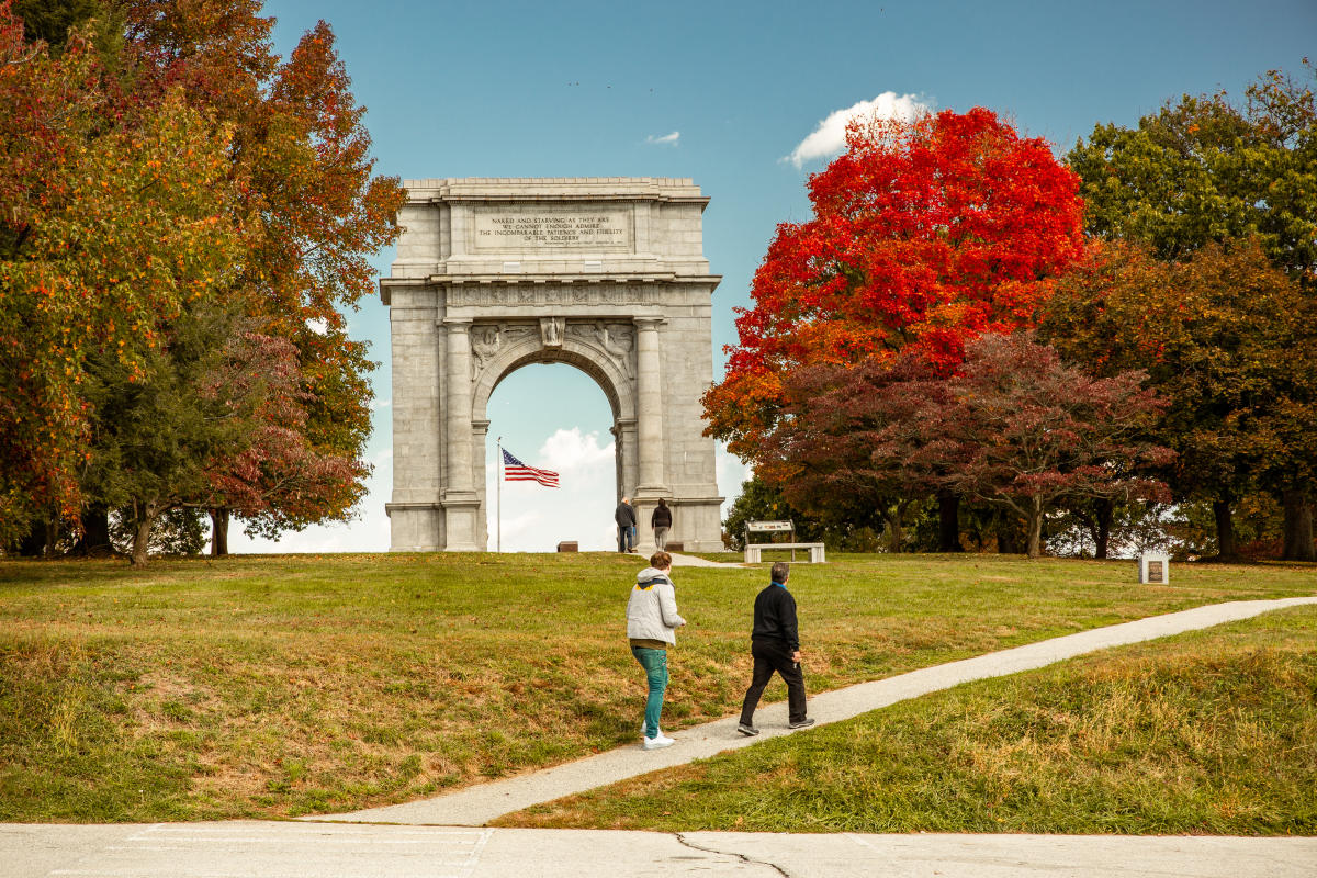 An older male and female walk casually up a hill in front of the monument arch at the Valley Forge National Historic Park surrounded by trees with fall foliage.
