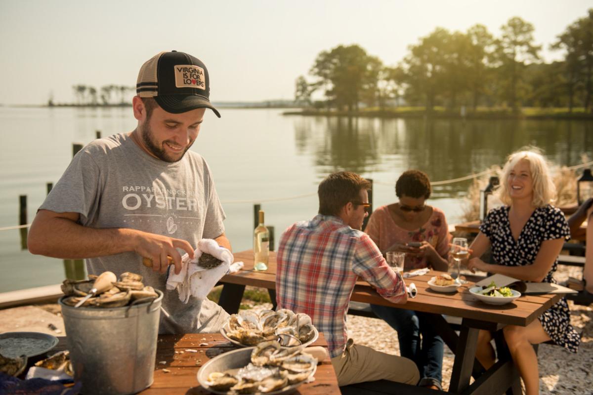 Merroir Restaurant employee shucks Rappahannock Oysters in front of a table of people dining on the waterfront