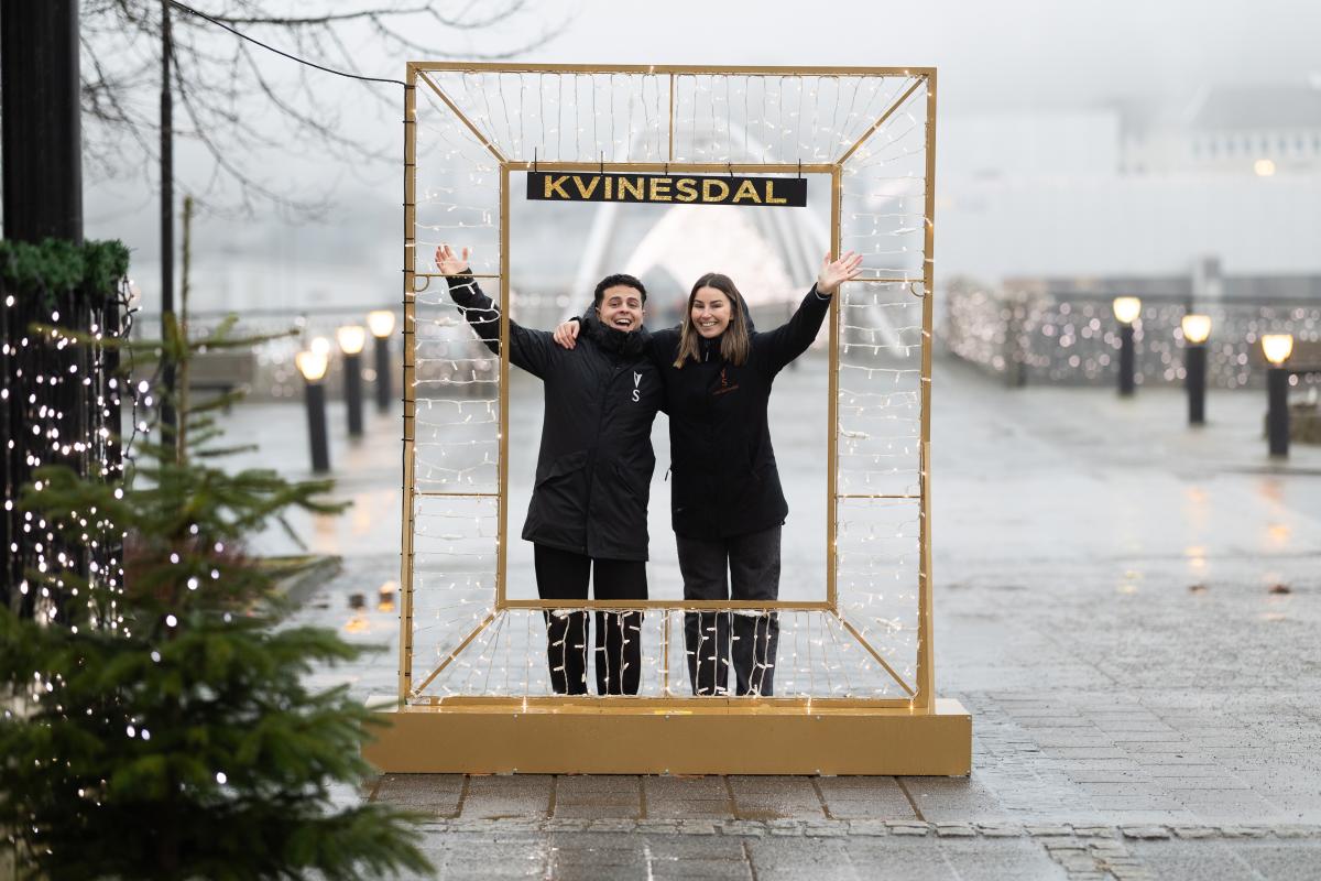 En mann og en dame står i en lysramme og vinker, over dem er det et skilt hvor det står "Kvinesdal"