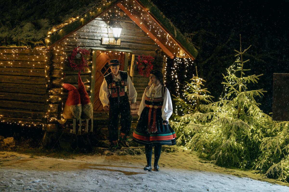 To bunadskledde kommer ut av et stabbur som er opplyst av julelys.