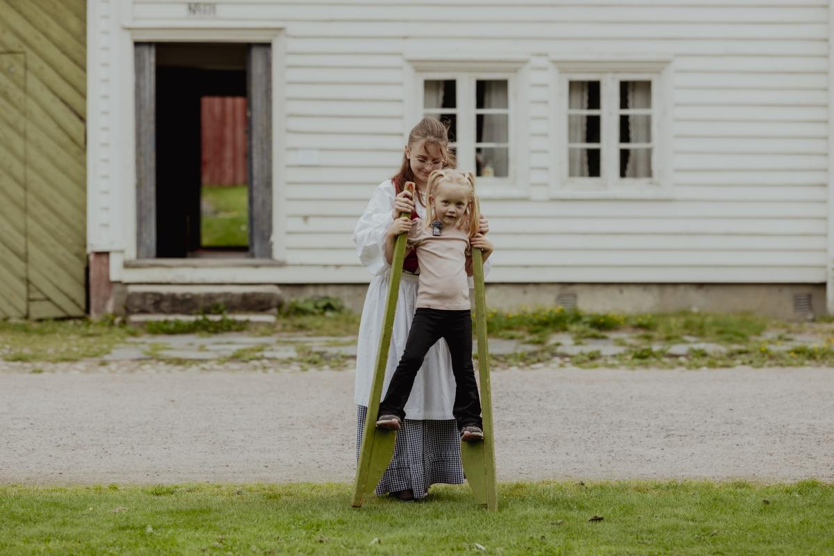 Jente prøver stylter foran gammeldagse hus, mens en kvinne i historiske klær hjelper henne