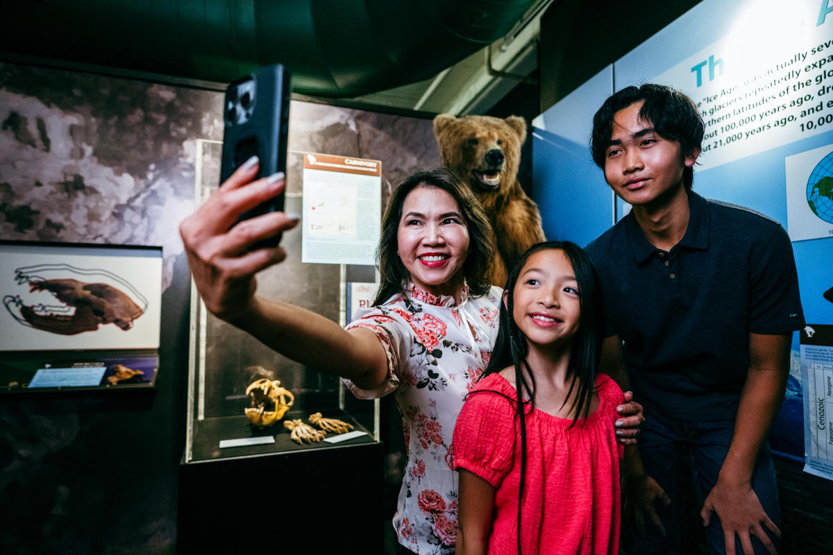 A mom and her son and daughter take a selfie in front of a stuffed Grizzly Bear at Exploration Place.