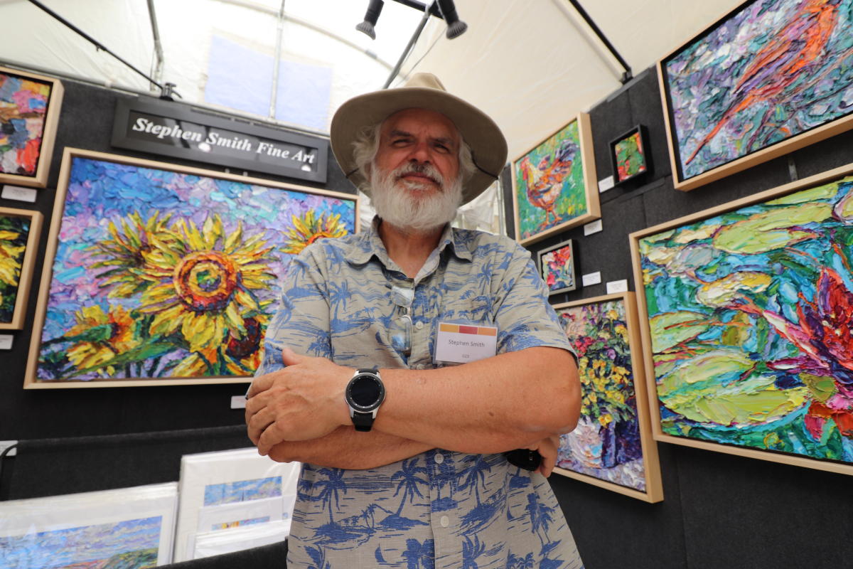 An artist poses in front of his paintings at his booth at Autumn & Art