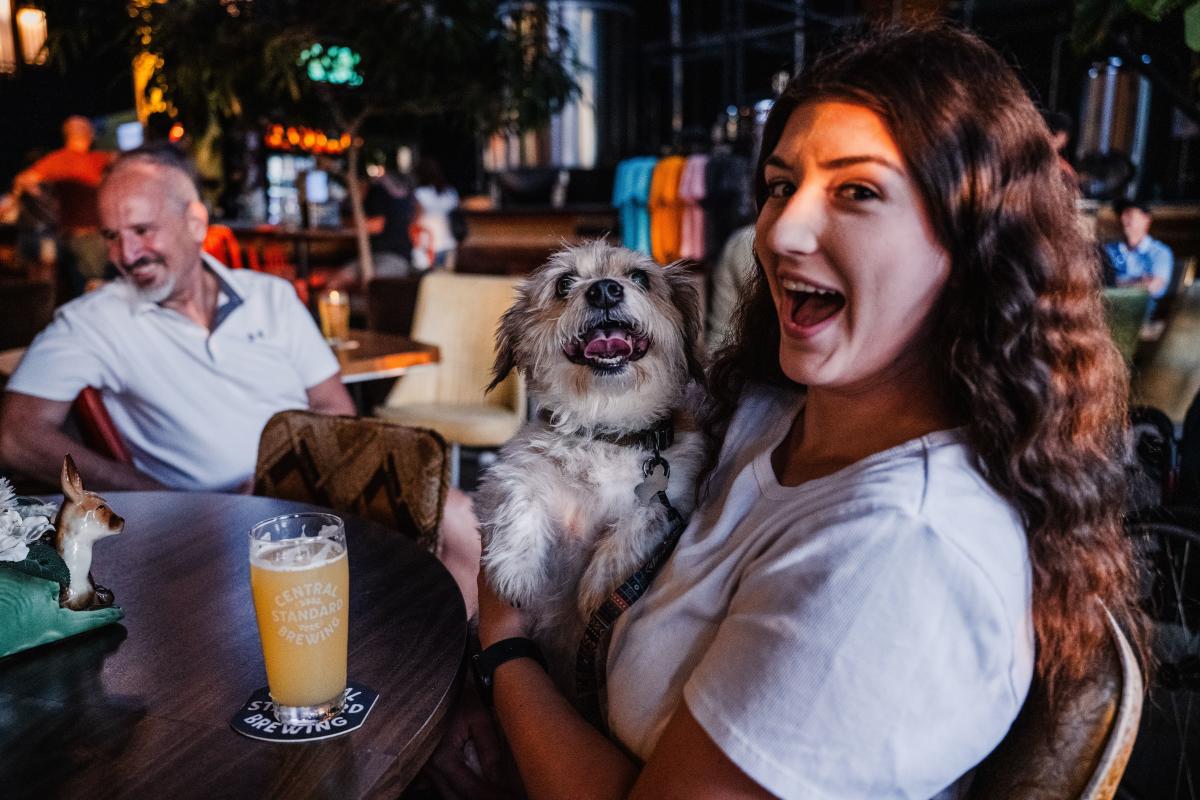 A young woman smiles while holding a dog and enjoying a craft beer at Central Standard Brewing.