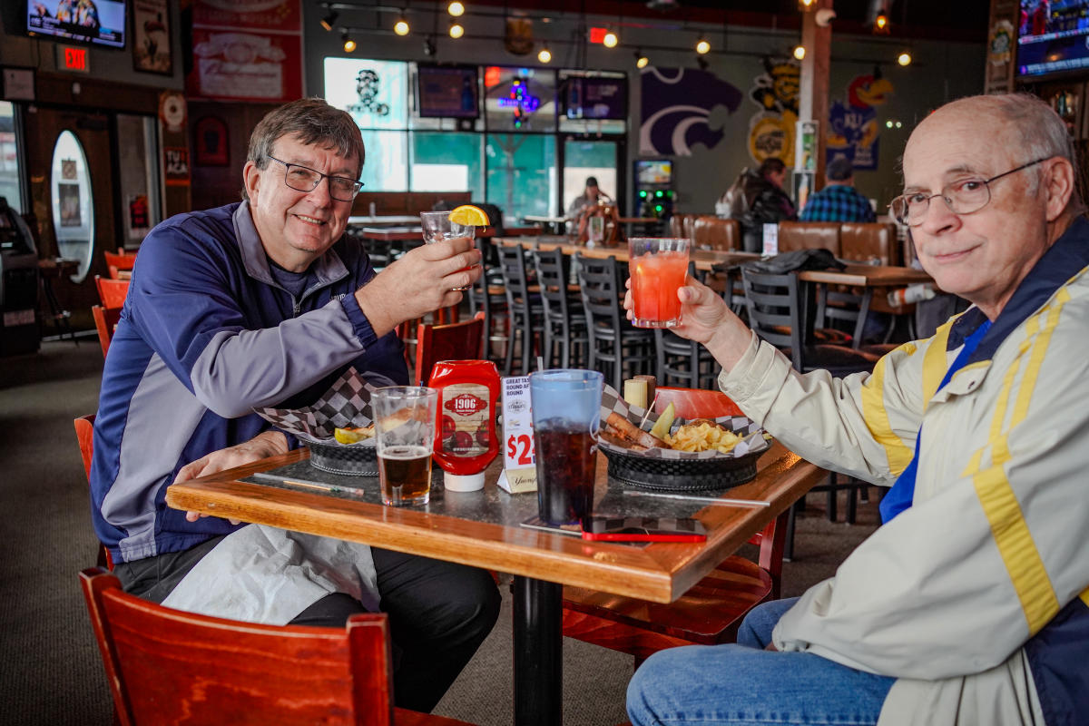 Two men raise their cocktails with Greatness Vodka at a table inside a bar
