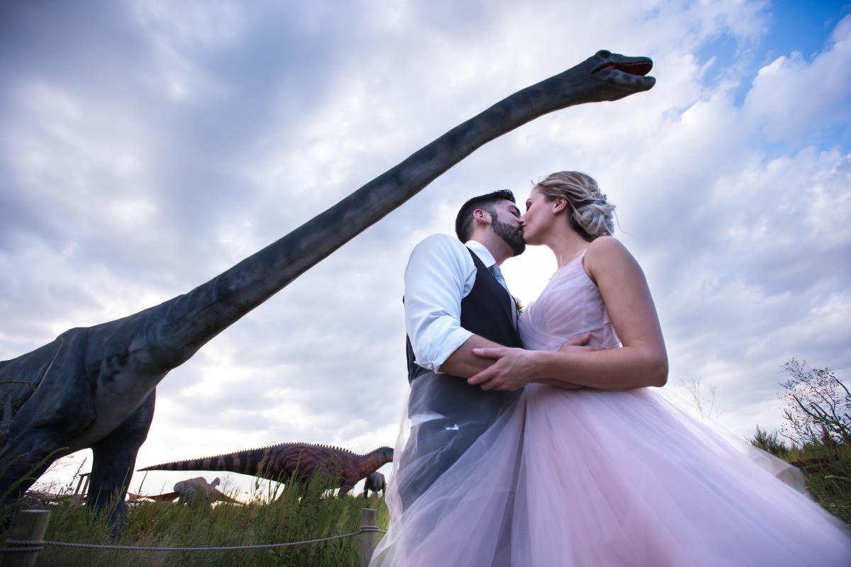 A bride and groom kiss beneath an animatronic dinosaur at Field Station: Dinosaurs