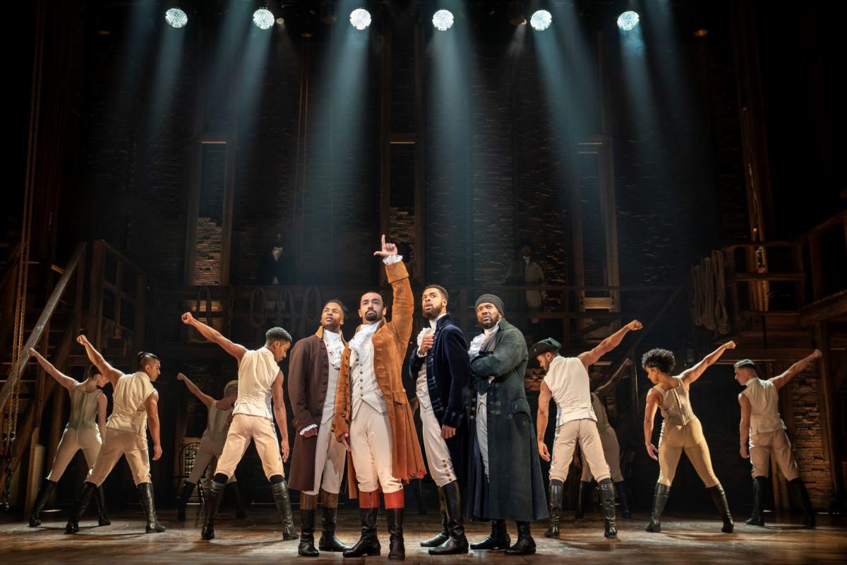 The cast of the musical Hamilton performs during a scene under bright lights