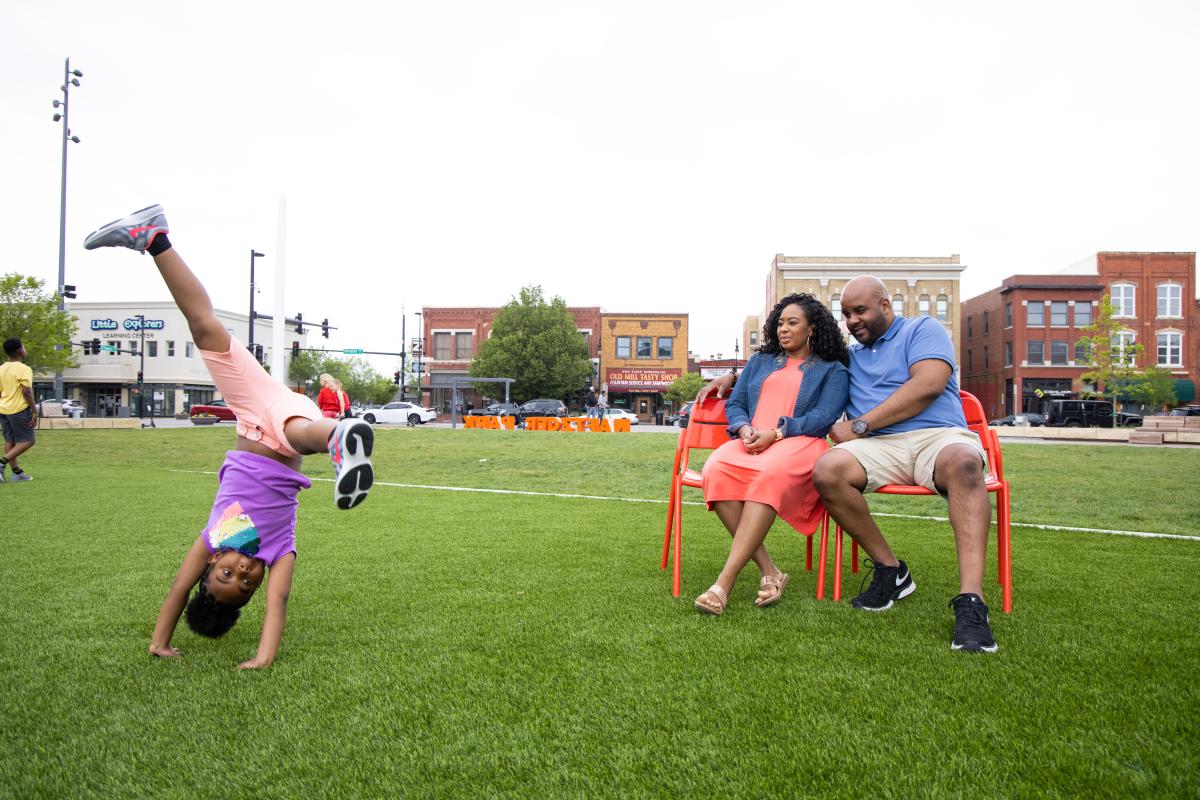A mother and father sit on a bench in Naftzger Park while their kids do cartwheels In Wichita, KS