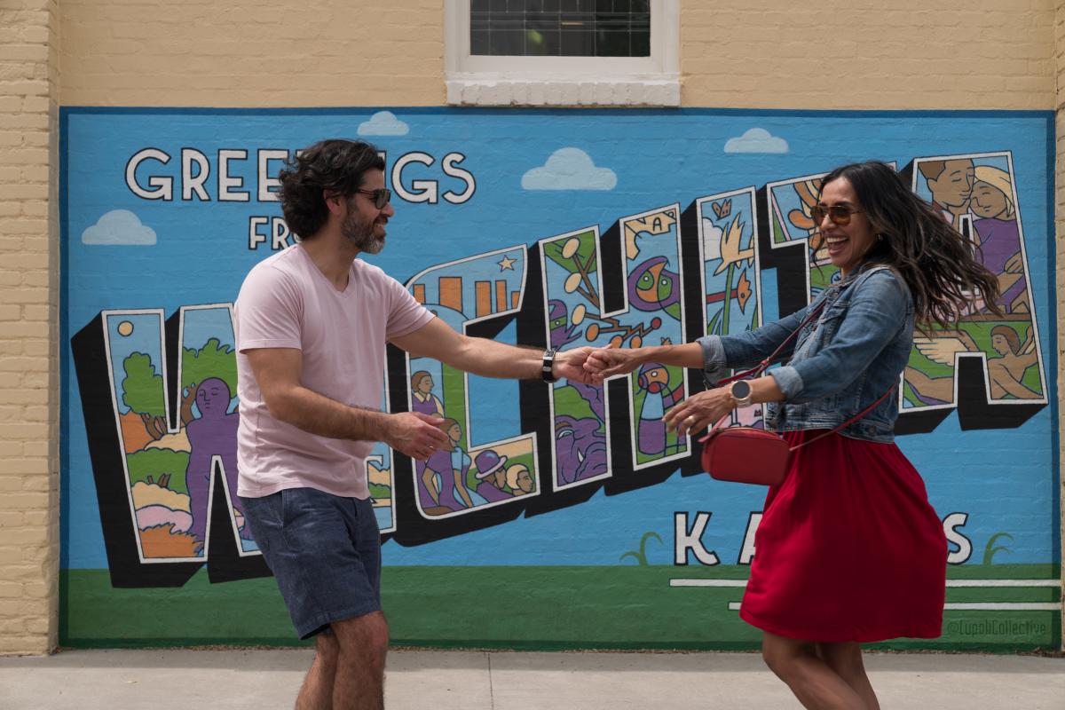 A Hispanic couple dances in front of a mural that says "Greetings from Wichita, Kansas