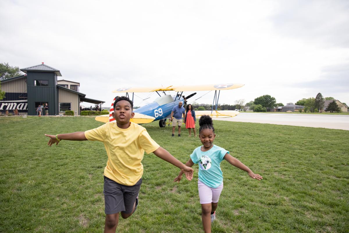 Kids run in a field by a plane at Stearman Airfield in Benton, Kansas