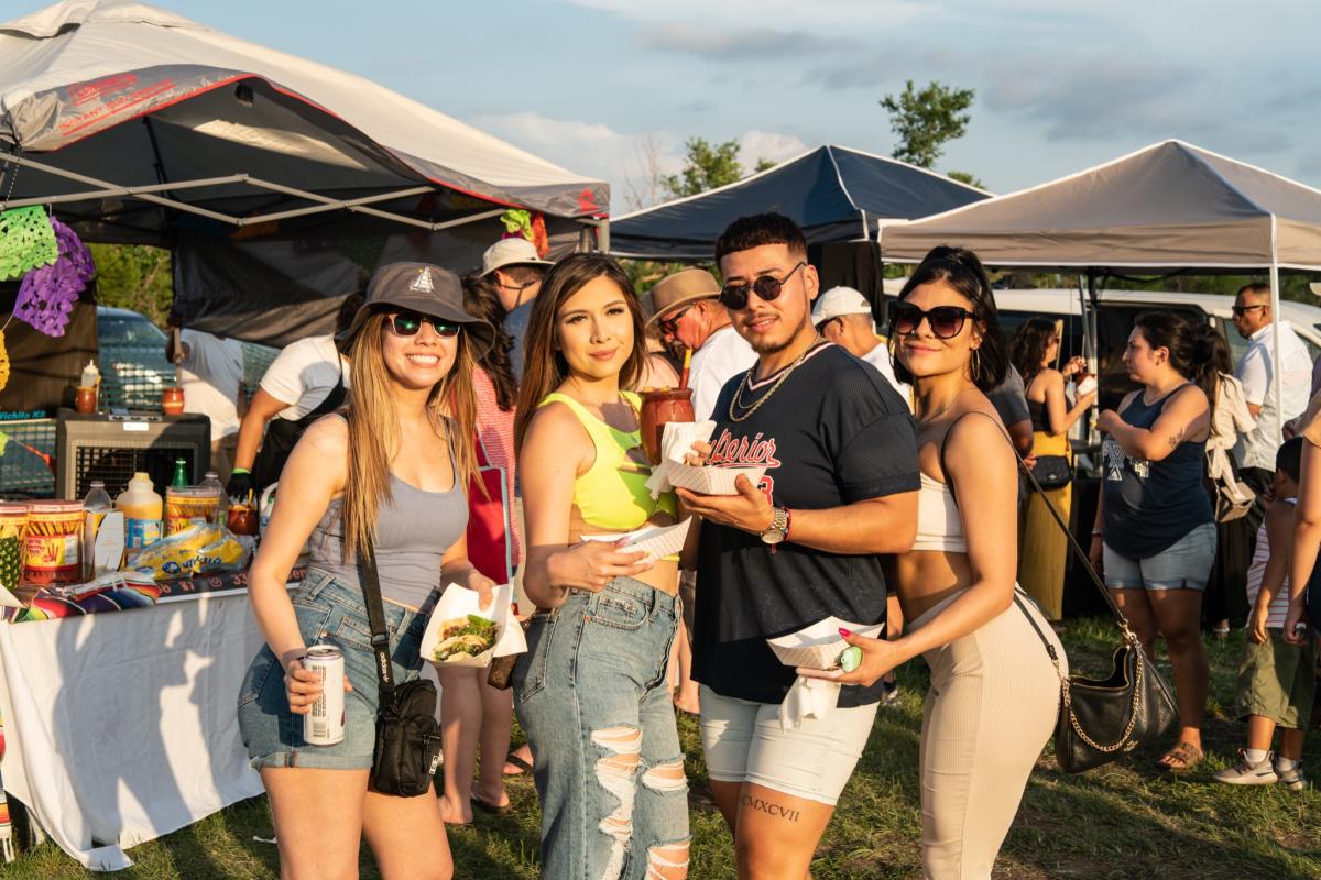 Four young adults gather in front of food vendor tents to take a photo at Wichita Taco Fest