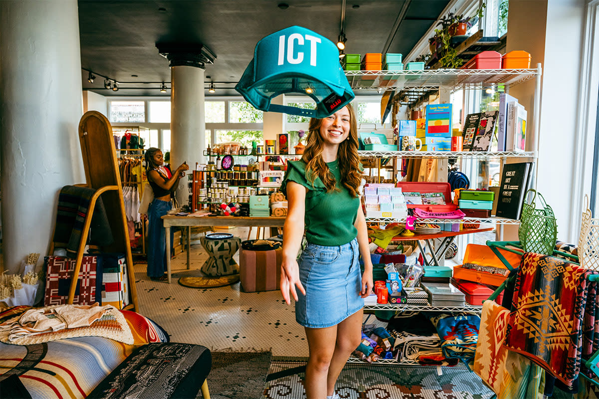 A young woman playfully tosses a hat that says "ICT" toward the camera while shopping at The Workroom