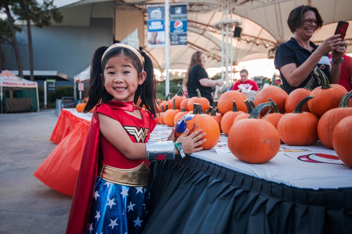 Little girl dressed up as Wonder Woman standing near a pumpkin during Hocus Pocus Pops at The Cynthia Woods Mitchell Pavilion in The Woodlands, Texas