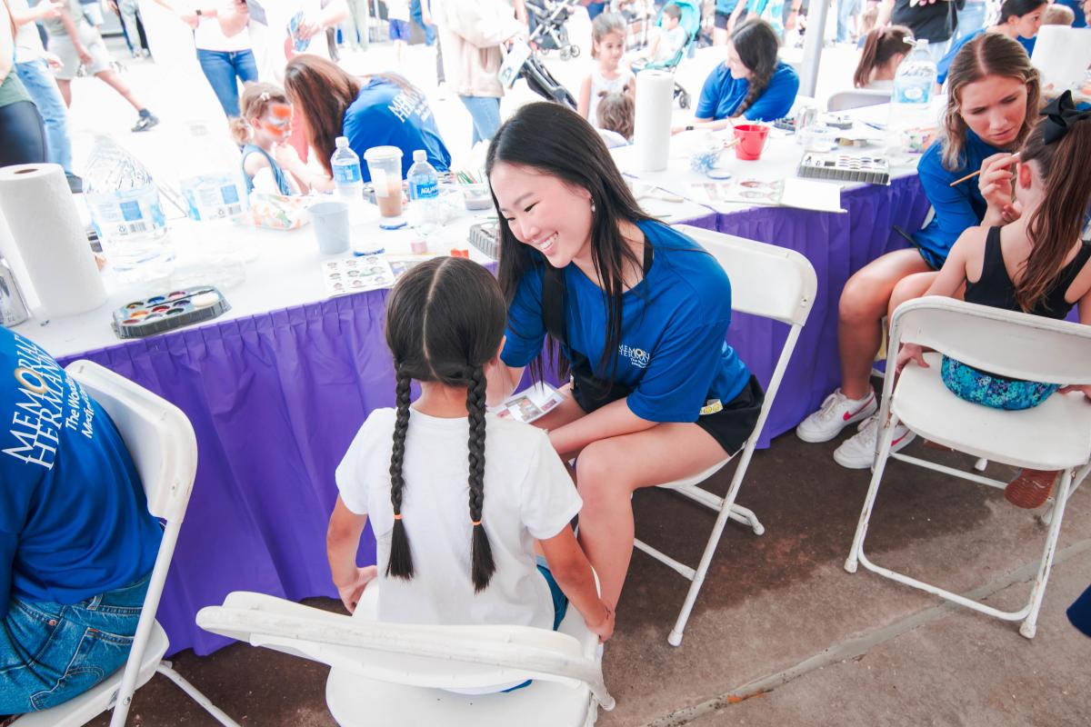 A young Asian woman smiles as she paints the face of a young Latina girl (facing away from the camera) with two long braids.