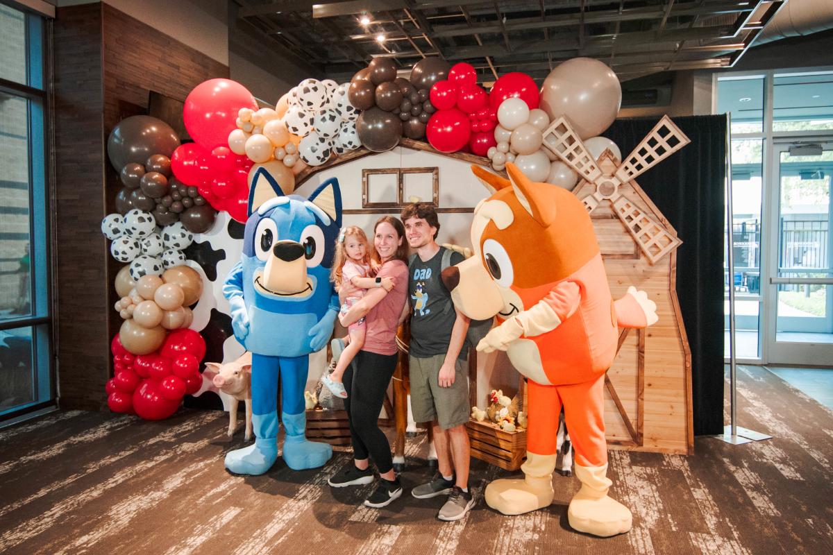 A young couple and their daughter (in the mother's arms) pose for a photo with a Bluey and Bingo from the children's TV show, "Bluey." Behind them is an elaborate wooden standee/backdrop of a barn and windmill surrounded by balloons in red, various shades of brown, and black and white cow patterns.