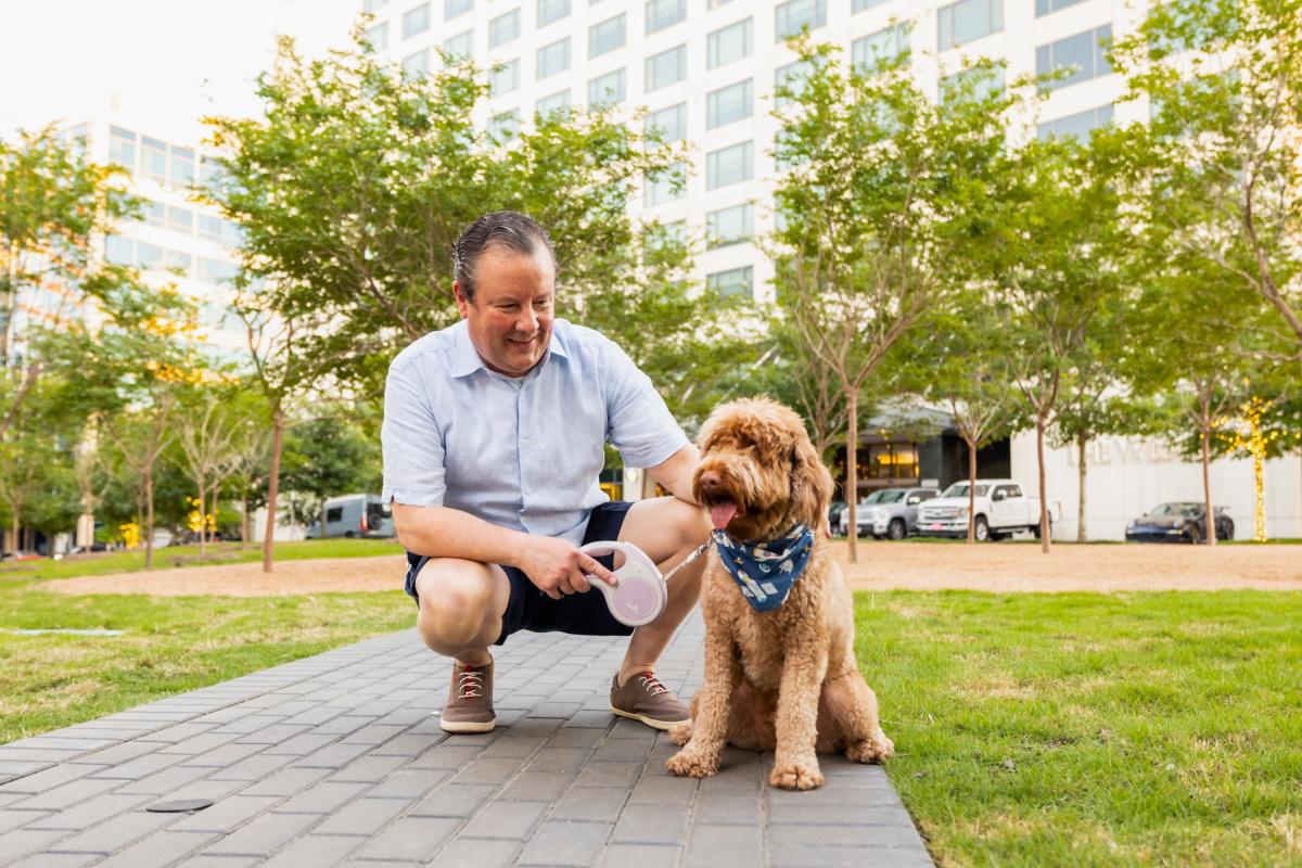 Nick Wolda, wearing a pale blue shirt and navy shorts, kneels down to pet his fluffy brown dog, Graham. The dog is wearing a blue bandana with rocket ship printing around his neck.