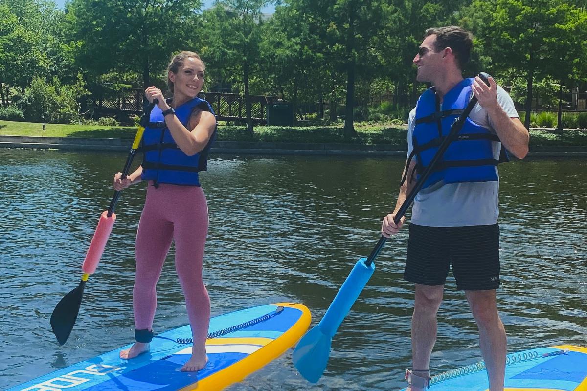 A young couple smiles at each other as they paddleboard down The Waterway toward Riva Row Boat House. The woman is wearing pink athletic pants and a bright blue life vest. The man is wearing black swim trunks, a white T-shirt, and a blue life vest. Both are holding paddles with a pink or blue pool noodle around one end.