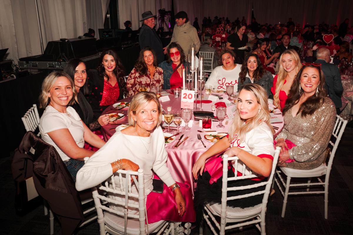 Eleven women dine at The Pavilion's "For The Love of the Arts" gala, including Linda Nelson in front. Some are dressed up, some are casual, but all are wearing themed attire in red, pink, and white.