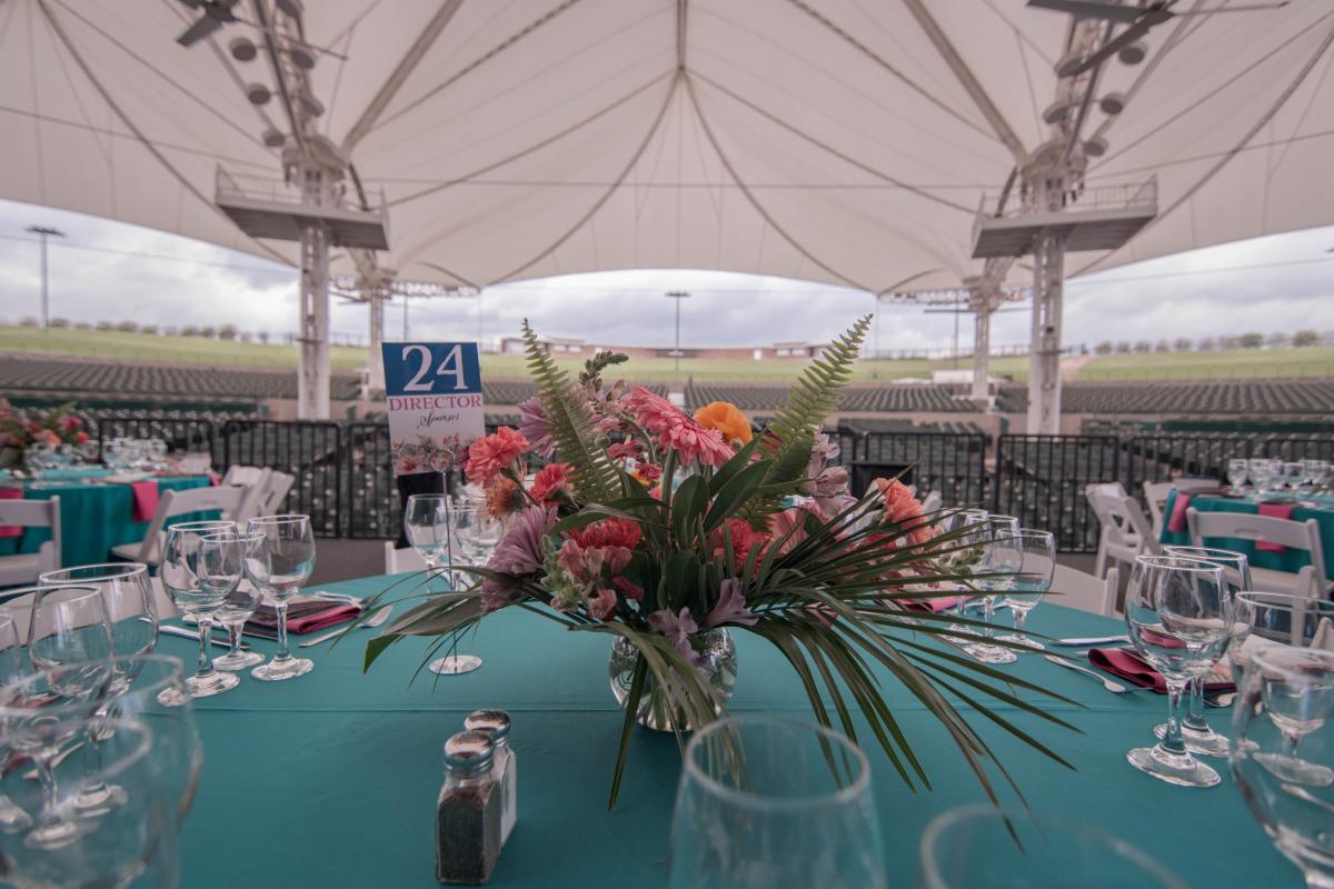 The camera focuses on a table set for The Cynthia Woods Mitchell Pavilion | Pavilion Partners Annual Wine Dinner and Auction. The table is under The Pavilion tent and covered with a dark teal cloth. It is set with empty wine glasses, red cloth napkins, and silverware. There is also a pretty floral centerpiece with fern fronds and pink and orange flowers.