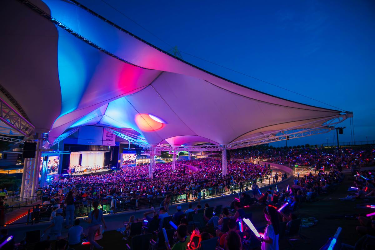 A wide-angle view of The Pavilion from the back of the lawn facing the left side of the stage. The entire audience is packed with people of all ages, many holding thick glowing wants. The underside of The Pavilion tents are lit up pink, blue, and purple.