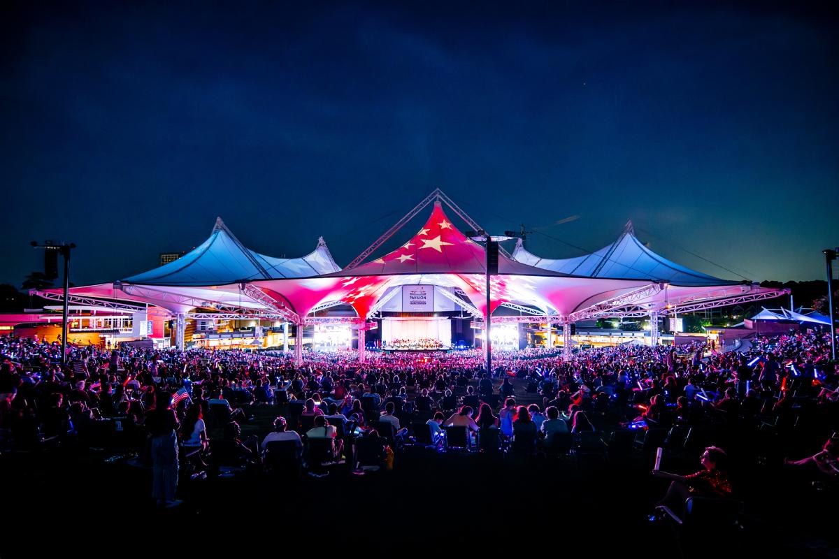 A wide-angle view of The Pavilion during the 2024 Star-Spangled Salute. The tents are lit up red, white, and blue with projections of stars across the red. The Houston Symphony is barely visible on stage, but every seat is packed from the front row to the back of the lawn. The sky is dark navy.