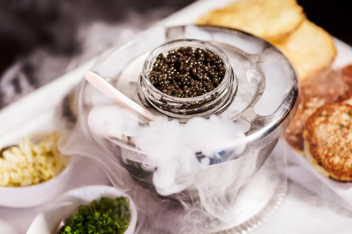 A metal dish featuring chilled caviar and accoutrements like bread slices, chives, and roasted garlic.
