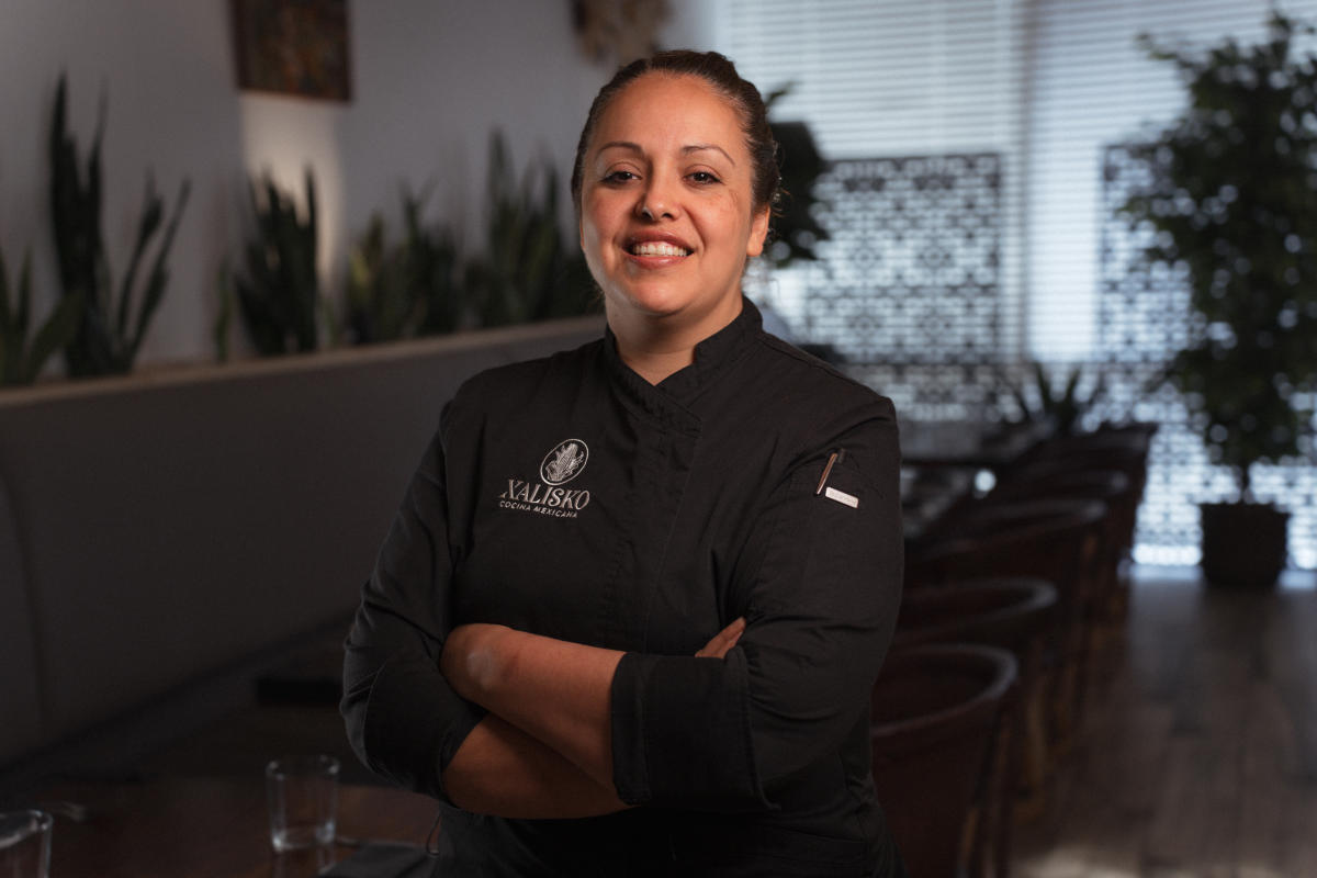 A Mexican woman (Chef Beatriz Martines, 30s-40s) smiles at the camera. She is wearing a black chef's uniform with a white logo of corn and white text that reads, "XALISKO COCINA MEXICANA."