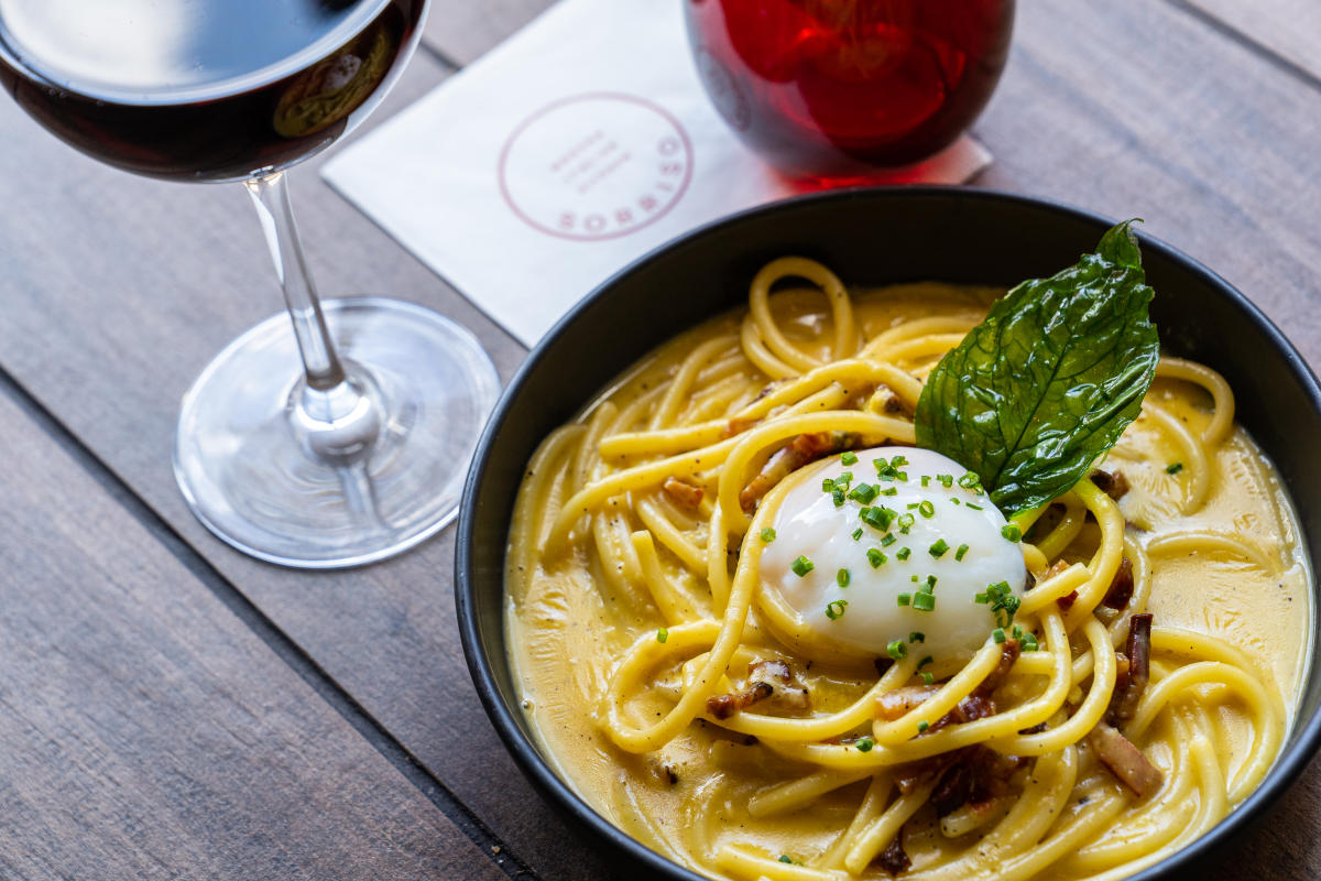 In the lower right corner of the frame is a black bowl of white pasta topped with a melted cheese ball, chives, and a mint leaf. Above the bowl is a red glass for water, and to the left, a glass of red wine. The setting is on a patio table on an overcast day.