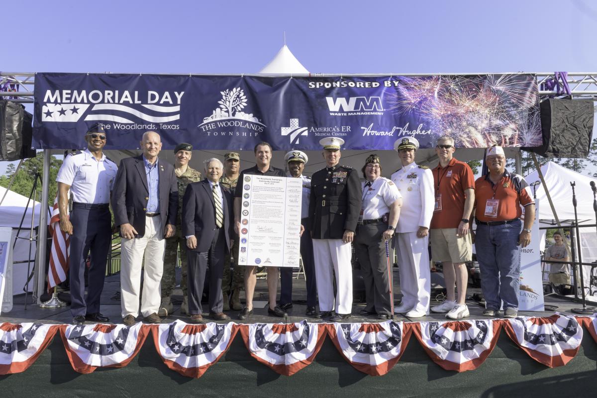 A group of twelve U.S. government and military leaders (men and women) stand on the stage, smiling at the camera. The man in the middle is holding a posterboard titled, "The Woodlands Township's Community Covenant," and bears the seals of each branch of the military.