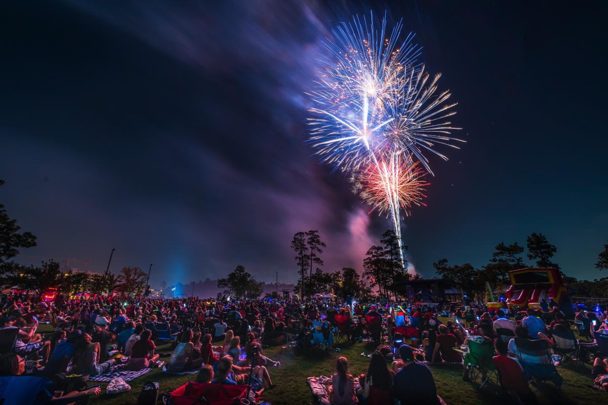 A bouquet of red, white, and blue fireworks burst in the night sky over Town Green Park. Hundreds of spectators stare up at the spectacle. Along the horizon are dark silhouettes of trees and a large, inflatable water slide.