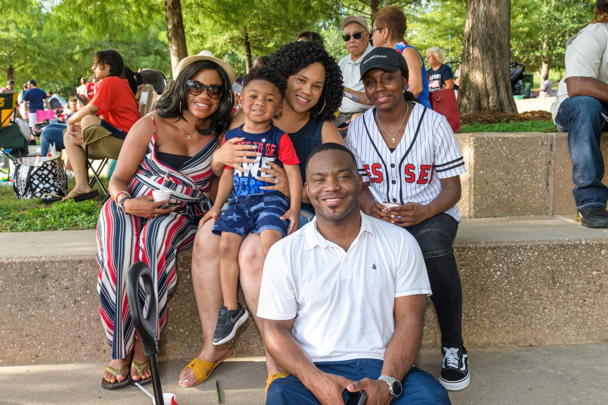 A family of five (seemingly a couple and their son, plus two other women who could be sisters to one of the parents) smile at the camera. They're sitting on outdoor steps, dressed all in red, white, and blue, enjoying the good weather and great Independence Day festivities at Town Green Park.