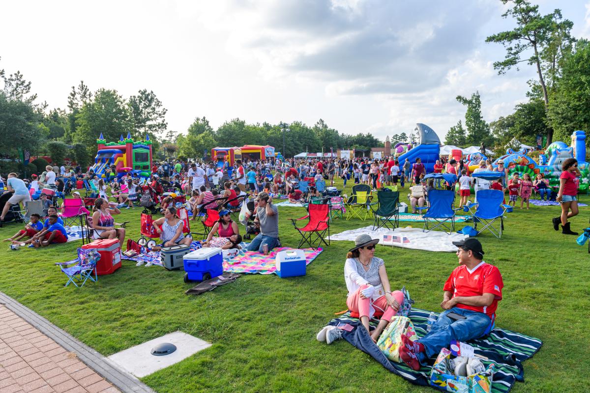 The camera looks out over Town Green Park, crowded with bounce houses, lawn chairs, picnic blankets, and people wearing patriotic colors, all in celebration of the July 4th Red, Hot, & Blue Festival.