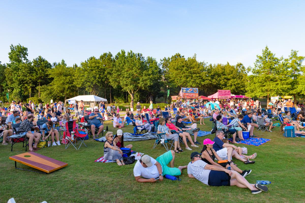 A crowd dressed in red, white, and blue relaxes in lawn chairs or on the grass in Town Green Park. On the left is a game of corn hole, and in the back right are various carnival food vendors and a snow cone truck.