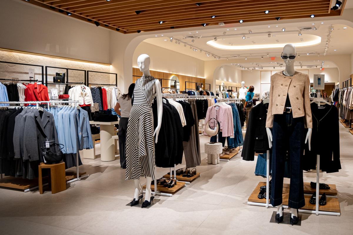 An overview of the Mango store in The Woodlands Mall. Alongside many racks of business coats, purses, and shoes for women, there are two mannequins standing front and center. The one on the left is wearing black heels with a classy, asymmetrical dress that has black and white stripes running in different directions. The other is wearing a tan business jacket with gold buttons, dark straight-leg pants, black heels, and black sunglasses.