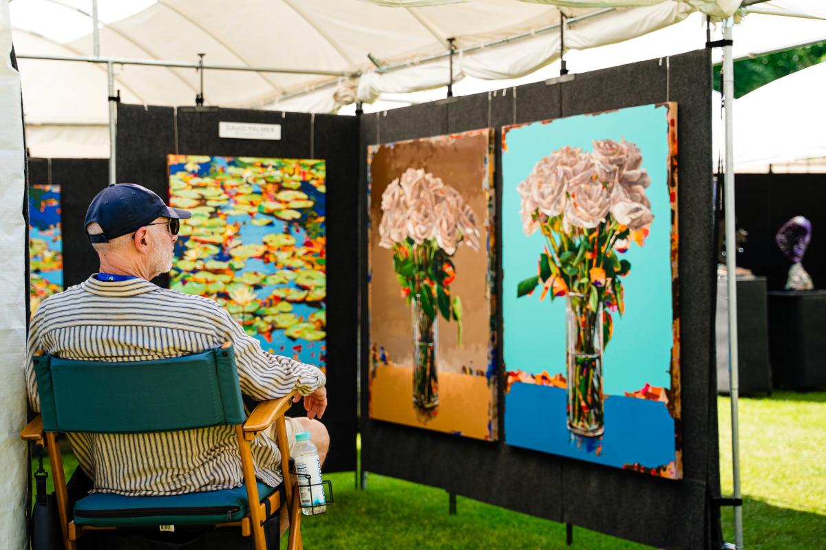 An artist sits in a director's chair facing his paintings. The man is wearing a striped shirt and a navy ballcap. Two of the paintings are of white roses on colorful backgrounds, another is of lily pads on the water.