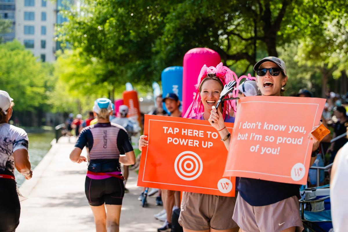 Two women cheer on IRONMAN athletes running along The Waterway. They're holding up signs by Visit The Woodlands that read, "TAP HERE TO POWER UP" and "I don't know you, but I'm so proud of you!"
