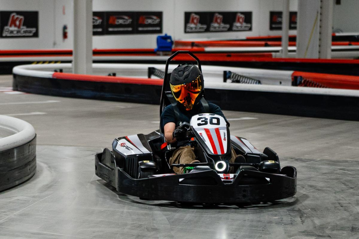 A man turns a go kart around a corner at K1 Speed. He's wearing a dark T-shirt, tan cargo shorts, black racing gloves, and a black motorcycle helmet with a colorful reflective visor. The track is gray and rubbery, the barriers alternating in red and white stripes. K1 posters line the back wall.