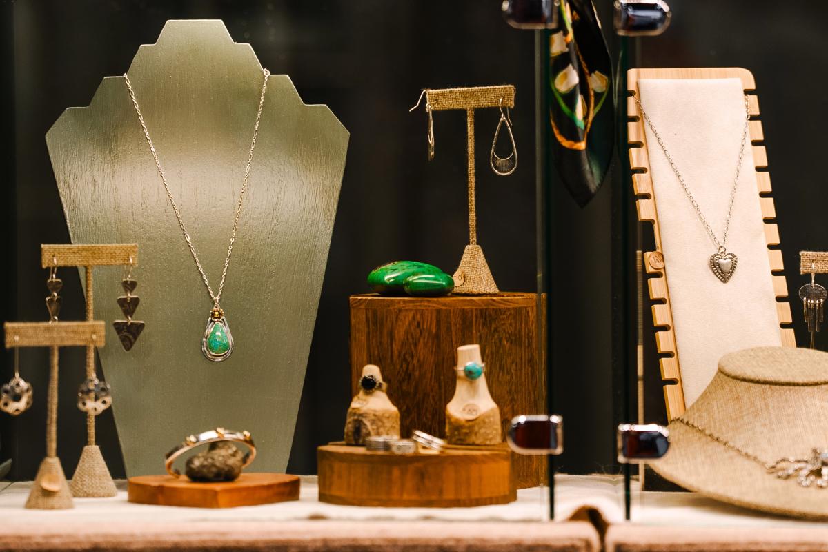 Close-up of jewelry displays at the Texas Fine Craft Show. The central piece is a chrysocolla teardrop-shaped pendant set in silver. Other stone and metal pieces are positioned strategically around it: pendants, rings, earrings, bracelet, and tumbled stone pieces.