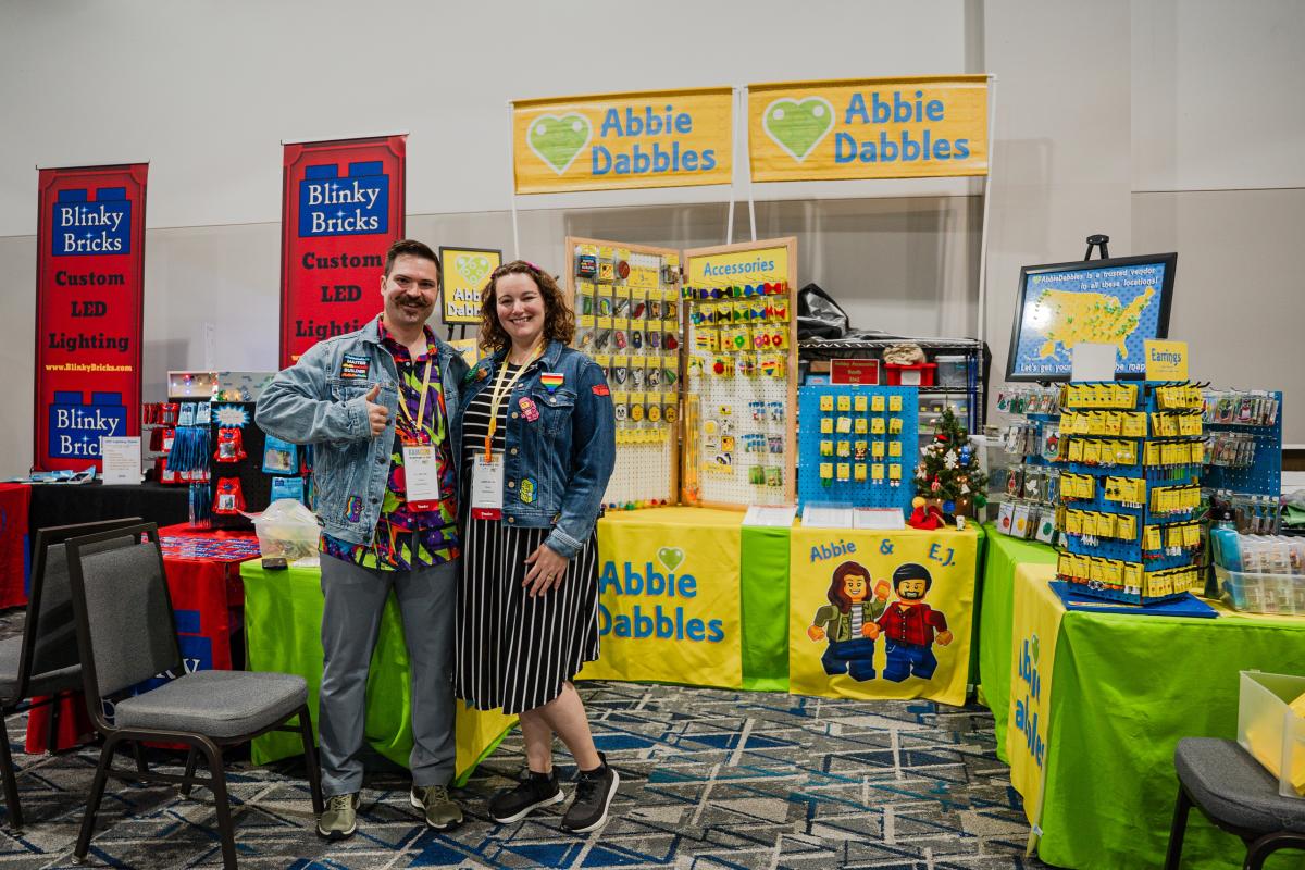 Married couple and owners of Abbie Dabbles bricks and minifigs accessories, Abbie and E.J., pose with their booth at BAMCOM, hosted at The Woodlands Waterway Marriott. E.J. has a curly mustache and is wearing a bright, multicolored shirt with a denim jacket bearing various patches he created. Abbie is wearing a black-and-white striped dress with a denim jacket like E.J.'s.