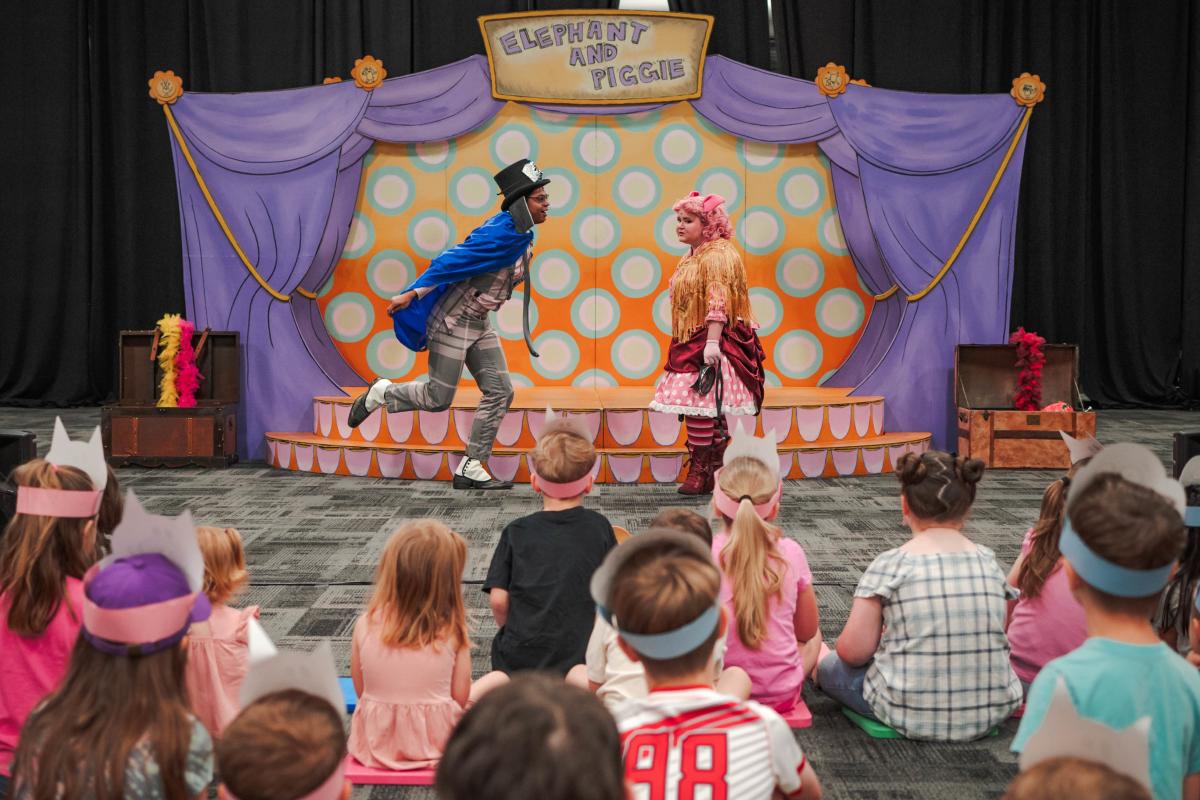 Two actors perform for children at The Pavilion. The male actor is dressed in gray plaid suit, blue cape, top hat, and elephant ears. The actress is wearing a curly pink wig, pig ears, and layered pink polka-dot skirts.