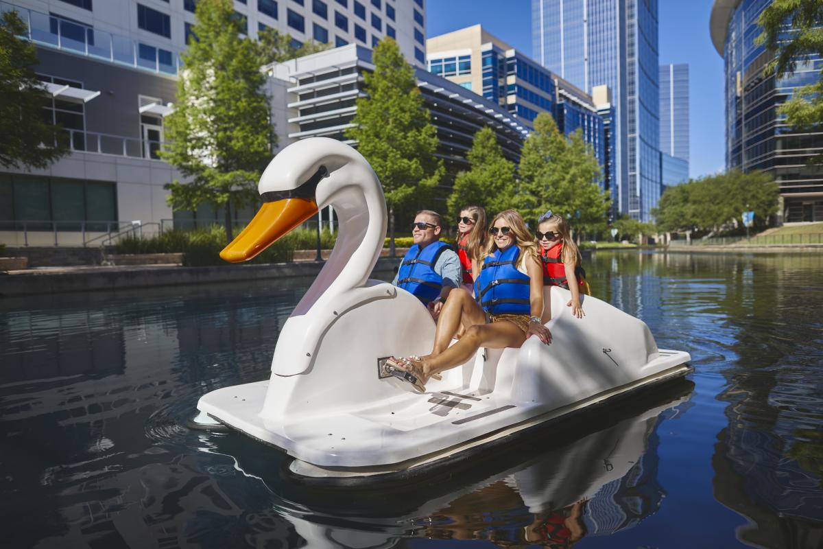 A family of four paddles a swan boat down The Woodlands Waterway. The parents (a white couple with blond hair and blue lifejackets) are sitting in the front of the boat. Two daughters (around 6-8 years old, wearing bright red-orange lifejackets) are standing in the back of the boat, leaning in over their parents' shoulders. Behind them are lush green trees and modern office buildings with windows reflecting the bright blue sky.