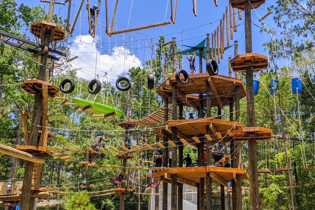 The camera looks up at the Texas TreeVentures aerial obstacle course. It is constructed of elevated wood platforms, like a treehouse, with various ropes, ladders, tire swings, and blue barrels interconnecting them. The trees surrounding the course are lush and green.