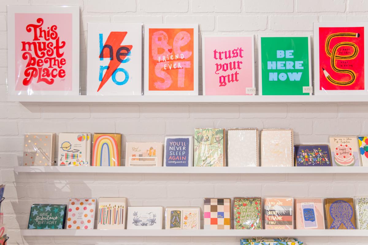 A wall of three white shelves, each lined with colorful greeting cards of various sizes.
