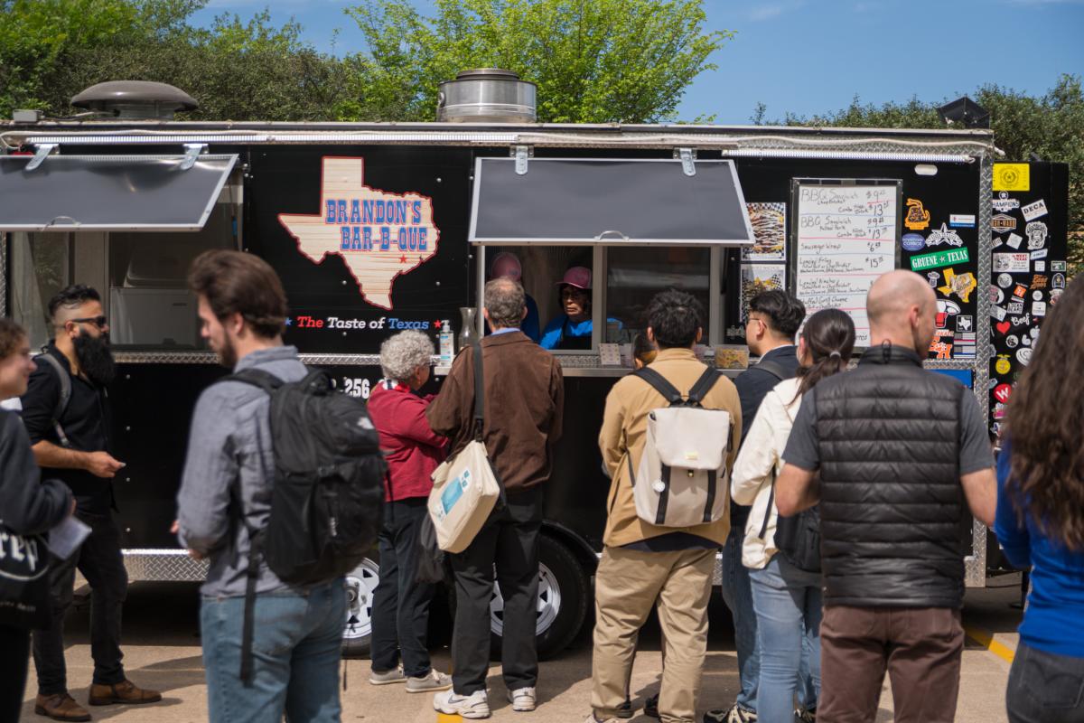 The camera faces the long side of a black food truck with two sliding windows, both open for business to a crowd in comfortable clothes. Between the two windows is a large, wood-textured, Texas-shaped logo with bright blue lettering that reads, "BRANDON'S BAR-B-QUE." On the far right, toward the front of the truck, is a white board menu and various stickers advertising Conroe, TX.